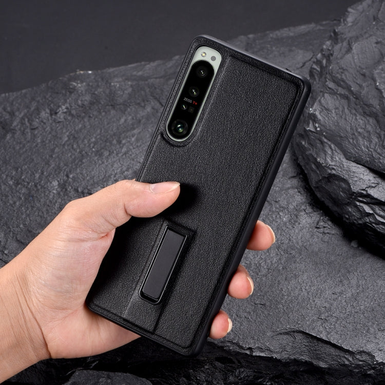 Black leather-texture protective phone case held in hand, showing camera cutout and holder.